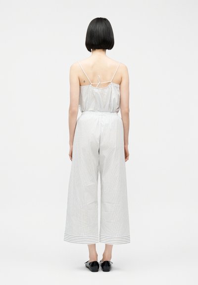 Γυναίκα με κοντά μαύρα μαλλιά, που φοράει ένα αμάνικο ολόσωμο σορτς με ανοιχτές ρίγες και φαρδιά πόδια, καθώς και μαύρα επίπεδα παπούτσια, στέκεται με την πλάτη γυρισμένη σε λευκό φόντο.