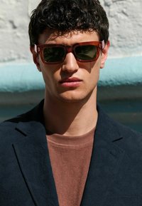 Skildpaddedesign solbriller med grønne linser; iført en struktureret marineblå blazer over en brun T-shirt med rund hals. Nærbillede af ansigt.