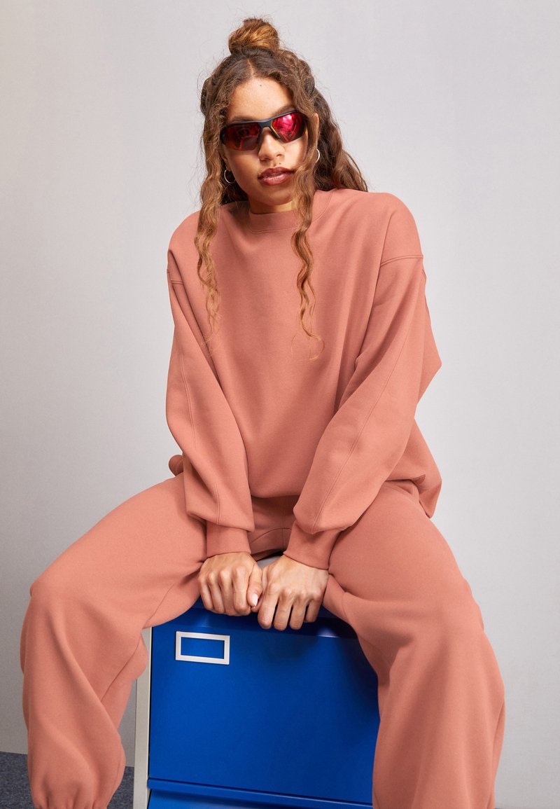 Sudadera rosa de gran tamaño y pantalones a juego hechos de tela suave. Persona sentada en un archivador azul, usando gafas de sol con tinte rojo.