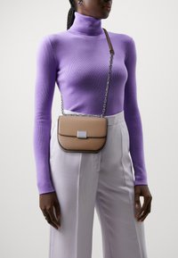 Brun crossbody-veske med klaff, sølv detaljer og kjede-stropp, båret over en lilla høyhalset genser og hvite vide bukser.