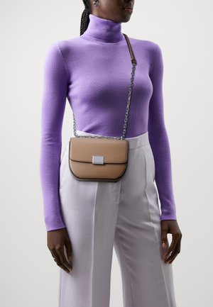 Borsa a tracolla color cuoio con pattina, accessori in argento e tracolla a catena, indossata su un dolcevita viola e pantaloni bianchi a gamba ampia.