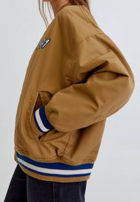 Widok z boku osoby w tanowym bomberze z niebieskimi i białymi paskami na mankietach i dolnym krawędzie, ręka w kieszeni, w połączeniu z ciemnymi spodniami.