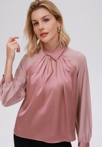 Blusa rosa chiaro in satin con colletto alla cinese, design frontale plissettato e maniche a palloncino. Texture liscia con una silhouette fluida.