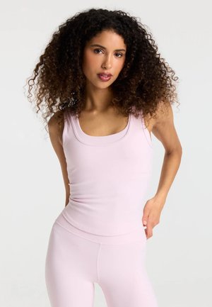 Kvinde med krøllet hår iført en lyserød ærmeløs top og matchende leggings mod en ensfarvet hvid baggrund.