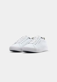 Par med vita låga sneakers med snörning, prydda med Tommy Hilfiger-märke på sidan och tungan, mot en helt vit bakgrund.