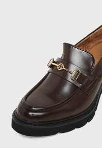 Mocassin en cuir marron foncé avec semelle noire épaisse et détail en métal doré en forme de mors de cheval sur l'avant.