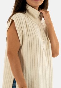 Donna che indossa un maglione senza maniche a coste di colore crema con un alto colletto e una cerniera corta sul davanti, toccando il colletto con una mano.