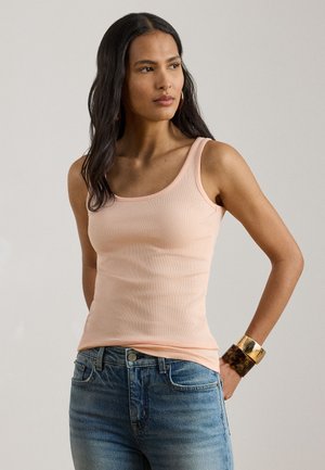 Pikkade tumedate juustega naine, kes kannab heleroosat varrukateta topi, siniseid teksapükse ja suuri kuldse ning kilpkonnakooreniitidest käevõrusid.