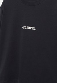 Czarna koszulka z białym napisem na środku klatki piersiowej: "ZBYT WRAŻLIWY NA WSPÓŁCZESNE CZASY" zapisaną wielkimi literami.