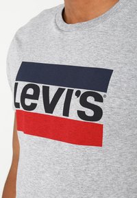 Camiseta de algodón gris con un diseño de rayas horizontales en azul marino y rojo, con "Levi's" destacado en letras negras en negrita.