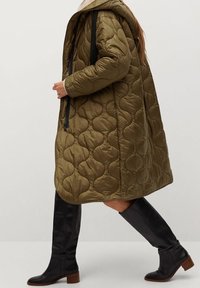 Olivengrøn quiltet frakke med hætte, der har runde mønstre, sort lynlås og kontrasterende sorte stropper. Knælange sorte støvler med brune hæle.