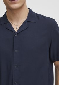 Donkerblauw shirt met korte mouwen en een knoopsluiting, met een gladde textuur en kleine knopen aan de voorkant. Een zilveren hangerketting is zichtbaar.