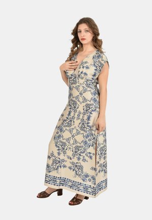 Donna con abito lungo beige con motivi floreali blu e spacco laterale, indossa tacchi aperti marroni, in piedi con una mano sul petto.