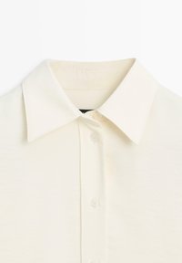 Camicia color crema con colletto appuntito, tessuto morbido e bottoni visibili lungo il davanti. Tessuto liscio con una leggera lucentezza.