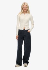 Kremowy prążkowany kardigan z krótkim dołem i guzikami, w zestawieniu z szerokimi, ciemnoniebieskimi spodniami z korduroy i jasnymi butami.