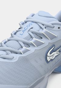 Αθλητικά παπούτσια ανοιχτού μπλε με υφασμάτινη επάνω επιφάνεια με υφή πλέγματος, που διαθέτουν λευκές λεπτομέρειες, λογότυπο Lacoste και ευέλικτους θηλιές κορδονιών.