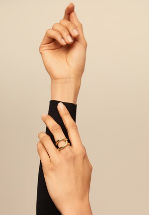 Anillo de tono dorado con diseño circular, acabado liso y detalle de gema incrustada, llevado en una mano sobre un fondo de manga negra.