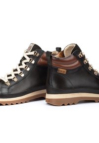 Botas de cuero negras de media caña con cordones crema, ojales metálicos, acentos marrones y una suela de goma con un patrón de relieve texturizado.