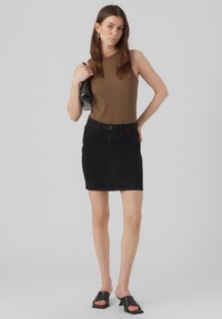 Débardeur marron à côtes accompagné d'une mini jupe en denim noir. Le mannequin tient un sac à main noir et porte des sandales à lanières noires. Fond gris uni.