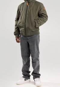 Olive zelená prošívaná bunda s předním zipem, žebrovanými manžetami a logovou nášivkou na rukávu, kombinovaná se šedými džíny a bílými teniskami.