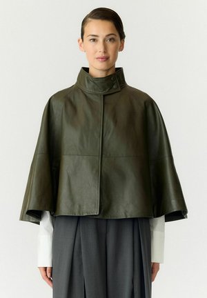 Modèle portant une veste en cuir vert olive foncé, de style cape, avec un col haut boutonné, par-dessus une chemise blanche et un pantalon gris anthracite.