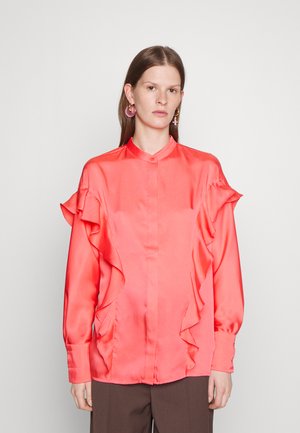 Γυναίκα με κοράλ σατέν μπλούζα με φούσκες κατά μήκος των μανικιών και μπροστά, συνδυασμένη με σκούρες καφέ παντελόνες και ροζ κρίκους στα αυτιά.