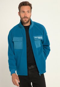 Giacca in pile teal con chiusura a zip, due tasche sul petto e tasche laterali. Presenta materiale a contrasto sulle tasche e dettagli di branding.