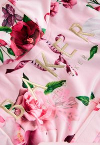 Tessuto rosa a fantasia floreale con vivaci rose e foglie. Testo ricamato in oro che dice "BAKER by TED". Texture morbida e aspetto caldo.