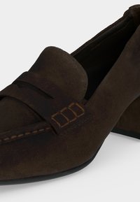 Mocassin en daim marron avec bride décorative et détails de piqûres, doté d'un petit talon bloc et d'un bout arrondi.