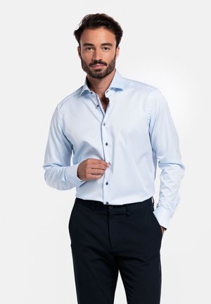 Lichtblauw overhemd met een spread collar en donkere knopen, gecombineerd met donkere broeken. Glad materiaal en nauwkeurige stikseldetails zijn zichtbaar.