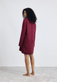 Vestito da notte lungo in cotone borgogna a maniche lunghe con righe verticali, dotato di colletto e polsini, indossato da una persona vista di spalle, a piedi nudi.