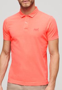 Heldere koraalkleurige polo met een zachte katoenstructuur, een klassieke kraag en een subtiel geborduurd logo op de borst. Korte mouwen.