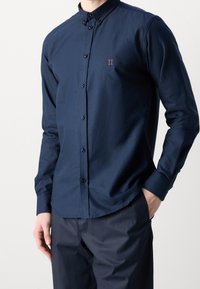 Mörkblå skjorta med knappar, krage och långa ärmar. Har en liten broderad "II" i orange på bröstet. Slät tygtextur.