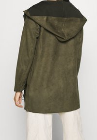 Olivgrön lång kappa med en kontrasterande svart huva, tillverkad av mjukt mikrofiberliknande material, utrustad med sidodragkedjor och en lös passform.