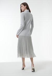 Vrouw in een lichtgrijze lange jurk met een geplooide rok en witte hoge hakken, staand en met de rug naar de camera gekeerd.