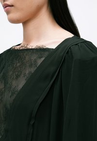 Mørkegrøn kjole med en struktureret blondeudskæring og gennemsigtig overlay. Plisseret skulderdetalje forbedrer det overordnede design.