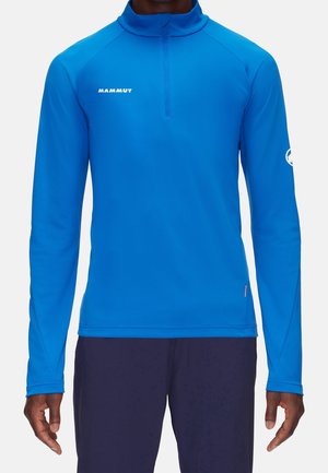 Homme portant un haut de sport à manches longues bleu vif avec une fermeture éclair quart, arborant le logo Mammut sur la poitrine et la manche, associé à un pantalon foncé.