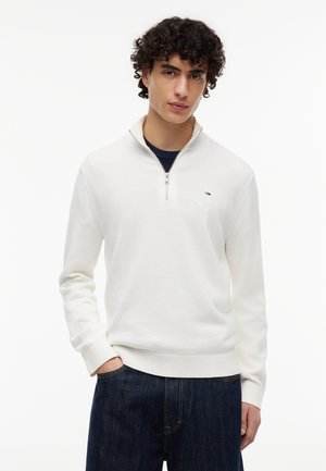 Junger Mann mit lockigem Haar, der einen weißen Pullover mit halbem Reißverschluss und dunkelblaue Jeans trägt, steht mit einer Hand in der Tasche vor weißem Hintergrund.