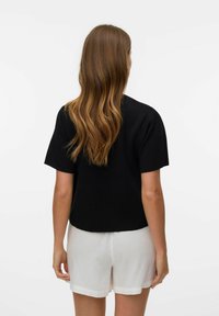 Kobieta z długimi falującymi brązowymi włosami ubrana jest w czarną bluzkę z krótkim rękawem i białe szorty, stoi tyłem na tle jednolitego białego tła.