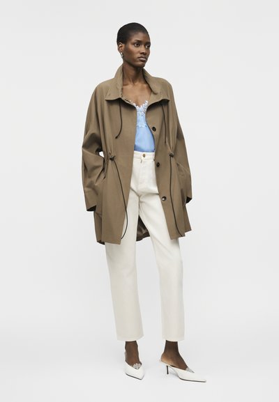 Žena oblečená do voľného hnedej kabáta cez svetlomodrý top s čipkou, biele vysoké nohavice a biele lodičky so špicatou špičkou.