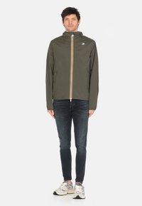 Giacca verde oliva con un alto colletto e una cerniera frontale, caratterizzata da un accento con strisce gialle; abbinata a jeans scuri e sneakers.