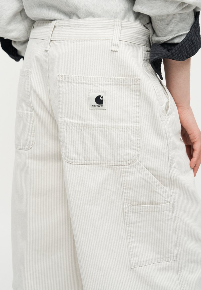 Pantaloni a righe beige chiaro con molte tasche, patch con logo Carhartt sulla tasca posteriore, indossati con camicia con maniche arrotolate e mano appoggiata sull'anca.
