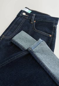 Tmavě modré džíny z denimu s ohrnutým lemem, viditelným švem, kovovým knoflíkem a přední kapsou na světlem pozadí.