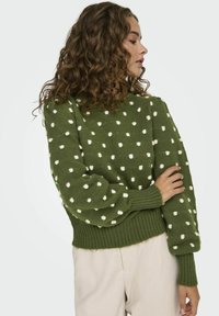 Groene trui met witte stippen, textuurgebreid, ronde halslijn, geribde manchetten en zoom; weergegeven op een model met krullend haar.