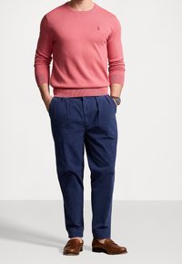 Ljusrosa stickad tröja med långa ärmar, marinblå veckade byxor och bruna skor. Har en liten logotyp på tröjan.