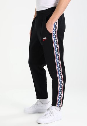 Mężczyzna ubrany w czarne spodnie dresowe Nike z niebiesko-białym wzorem na bocznych paskach, białe skarpetki i białe buty sportowe Nike, stojący z rękami w kieszeniach.