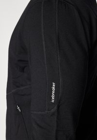Chemise noire à manches longues en tissu extensible, dotée de coutures plates, d'une poche zippée et du logo "icebreaker" sur la manche.
