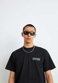 Svarte bomulls t-skjorter med en blå logo på venstre bryst, sammen med svarte solbriller med et moderne design og mørke linser. Sølvkjede.