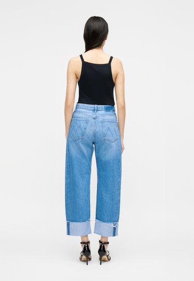 Γυναίκα που φοράει μαύρο φανελάκι, φαρδύ ανοιχτό μπλε τζιν με γυρισμένα μπατζάκια και μαύρα σανδάλια με ψηλά τακούνια, στέκεται με την πλάτη προς τον θεατή.