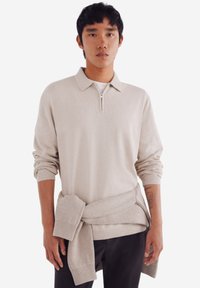 Beige Viertel-Reißverschluss-Pullover mit gerippten Bündchen und Saum. Das Modell trägt einen leichten Pullover, der an der Taille über einem weißen T-Shirt und einer dunklen Hose gebunden ist.
